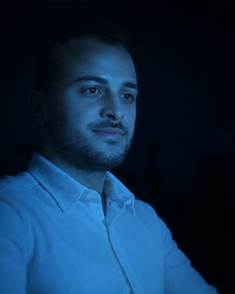 Enes Aktaş, lit from one side against a dark room — a frame from the film on the home page.
